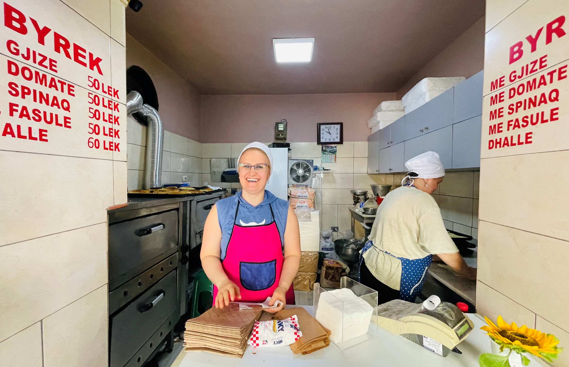 Smiling counter person with pink apron and a stack of paper bags waiting for an order